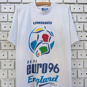 Puede incluir: Camiseta blanca con un logotipo Umbro azul y blanco, un gráfico de un jugador de fútbol y el texto "UEFA EURO 96 England".