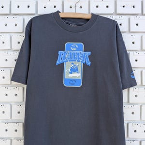 May include: Dark gray t-shirt with a blue and white graphic of a skateboarder inside a smartphone, with the word "HAWK" above. The shirt has short sleeves and a crew neck. The Hawk logo is also on the left sleeve.