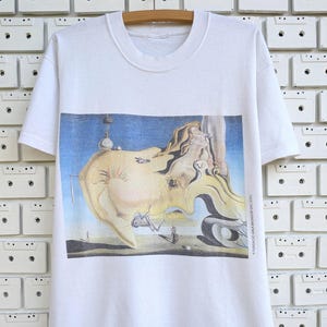 May include: White t-shirt featuring a surrealist painting with a landscape and distorted figures in shades of yellow, blue, and beige. The artwork is centered on the shirt, with the text "FUNDACIO GALA-SALVADOR DALI 2003" printed vertically on the right side.