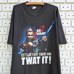 Puede incluir: Una camiseta negra con un estampado gráfico de un personaje del programa de televisión Red Dwarf. El personaje sostiene un arma y el texto "Let's get out there and TWAT IT!" está impreso debajo de la imagen.