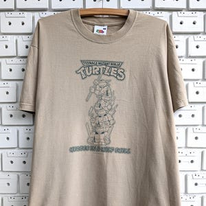Puede incluir: Camiseta color beige con el logo de las Tortugas Ninja y la frase "Heroes in a Half Shell". El gráfico muestra a las cuatro tortugas apiladas. La camiseta está colgada en una percha de madera.