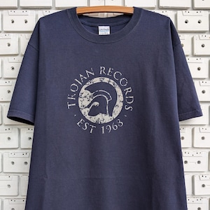 Könnte beinhalten: Ein marineblaues T-Shirt mit einem weißen, abgenutzten Druck des Trojan Records-Logos. Das Logo zeigt einen Kreis mit den Worten "Trojan Records" um den Außenrand und einen Helm innerhalb des Kreises. Die Worte "Est 1963" sind unter dem Kreis gedruckt.