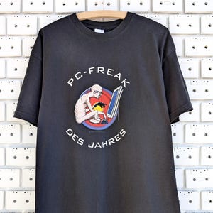 以下が含まれることがあります： 黒いTシャツ。座ってパソコンを操作する人物のグラフィックが描かれています。「PC-FREAK DES JAHRES」の文字が上下にアーチ状に配置されています。木製のハンガーにTシャツが掛けられています。