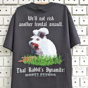 Op de afbeelding: Donkergrijs t-shirt met een afbeelding van een dreigend wit konijn met bebloede tanden en de tekst "We'll not risk another frontal assault." en "That Rabbit's Dynamite!" met het Monty Python-logo. Het konijn staat in groen gras.