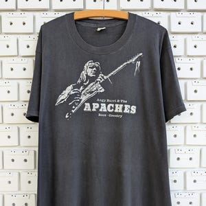 May include: Dark gray t-shirt featuring a white graphic of a person playing a guitar. The text "Angy Burri & The Apaches Rock-Country" is printed below the graphic. The shirt is hanging on a wooden hanger.
