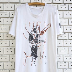 Puede incluir: Camiseta blanca con una ilustración gráfica en negro, marrón y rojo. El diseño presenta una figura sosteniendo un objeto, con el texto "Lola Blake" visible. La camiseta está colgada en una percha de madera.