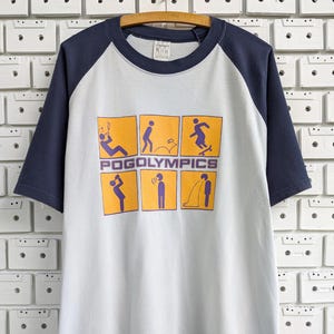 Puede incluir: Una camiseta raglán azul claro y azul marino con la palabra "POGOLYMPICS" en naranja y seis cuadrados naranjas con ilustraciones de figuras de palo. La camiseta está colgada en una percha de madera.