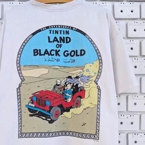 May include: White t-shirt with a graphic print of a cartoon jeep driving through a desert landscape. The graphic includes the text "The Adventures of Tintin Land of Black Gold".