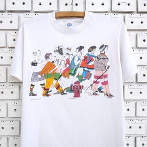 以下が含まれることがあります： カラフルな人々のグループが歩いているイラストが描かれた白いTシャツ。イラストは漫画風で、消火栓と犬が描かれています。