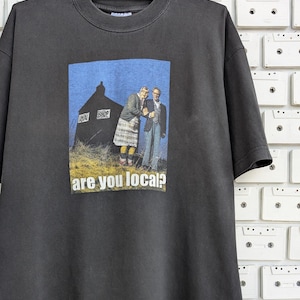 Könnte beinhalten: Ein dunkelgraues T-Shirt mit einem Grafikdruck von zwei Personen, die vor einem Gebäude mit dem Text "Local Shop" stehen. Der Text "are you local?" ist unter dem Grafikdruck gedruckt.
