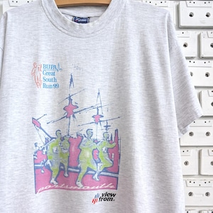 Op de afbeelding: Grijs T-shirt met een kleurrijke afbeelding van mensen die rennen. De afbeelding bevat de tekst "BUPA Great South Run 99" en "Portsmouth View from."