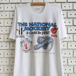 Pu&ograve; includere: Maglietta bianca con una stampa grafica blu e rossa di un calciatore. La grafica include il testo "The National Mockery It Could Be You!" e "Newcastle Five" e "Urinal Nil".