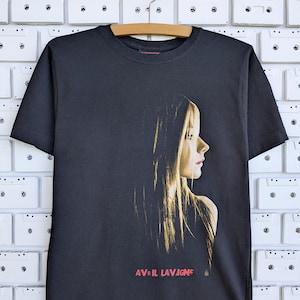 May include: A black short-sleeve t-shirt featuring a profile portrait of a person with long hair in golden tones. The name AVRIL LAVIGNE is printed in red text below the portrait. The shirt has a crew neckline and is displayed on a wooden hanger.