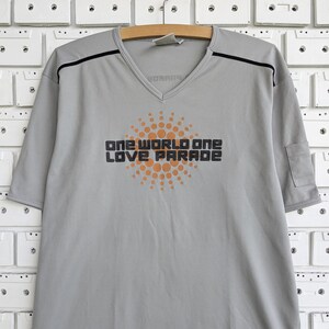May include: A light gray V-neck t-shirt featuring short sleeves and a small pocket on the left sleeve. The chest displays an orange circular sun graphic with the black text "ONE WORLD ONE LOVE PARADE" centered over it. Black piping accents the shoulders.