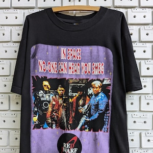 Op de afbeelding: Zwart T-shirt met een paarse afbeelding van de Red Dwarf-crew. De afbeelding bevat de tekst "In Space No-One Can Hear You Smeg" en het Red Dwarf-logo.
