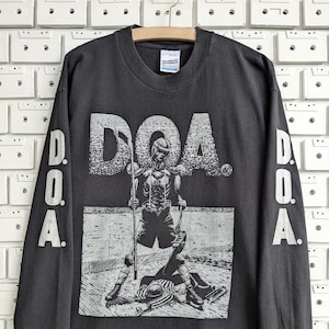 May include: A charcoal gray long-sleeve t-shirt featuring a graphic print of a hockey player and the text D.O.A. on the front. The sleeves also display the text D.O.A. printed vertically in a light gray color. The shirt has a crew neckline.