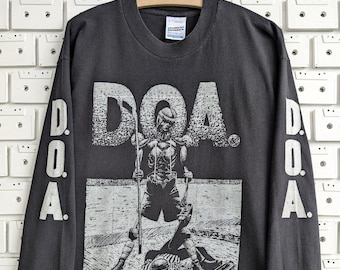 May include: A charcoal gray long-sleeve t-shirt featuring a graphic print of a hockey player and the text D.O.A. on the front. The sleeves also display the text D.O.A. printed vertically in a light gray color. The shirt has a crew neckline.