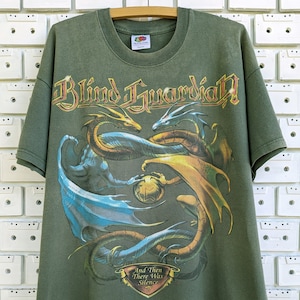 Op de afbeelding: Een groen T-shirt met een afbeelding van drie draken die om een gouden bol heen kronkelen. De bandnaam "Blind Guardian" is in gouden letters boven de draken gedrukt. De tekst "And Then There Was Silence" is onder de draken gedrukt.