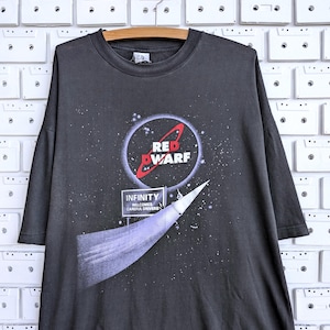 Puede incluir: Una camiseta gris carbón de manga corta con un gráfico del logotipo de Red Dwarf dentro de una escena espacial circular. Debajo del logo hay una señal de tráfico que dice INFINITY WELCOMES CAREFUL DRIVERS, con un rastro de luz en la tela.