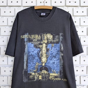Könnte beinhalten: Ein anthrazitfarbenes Kurzarm-T-Shirt mit einem Grafikdruck des Sepultura Chaos A.D. Albumcovers. Das Design zeigt eine zentrale Figur, die in einer mechanischen Struktur vor einem blauen, texturierten Hintergrund aufgehängt ist.