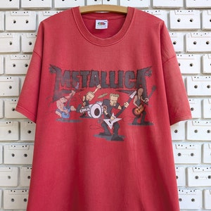 Puede incluir: Camiseta roja con una imagen de dibujos animados de la banda Metallica. Los miembros de la banda están tocando instrumentos y el nombre de la banda está escrito en negro encima de ellos.