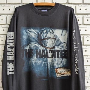 Puede incluir: Camiseta negra de manga larga con un estampado gráfico de una banda llamada "The Haunted". El gráfico presenta a una persona con la cabeza cubierta con un paño blanco y atado con una cuerda. El texto "one kill wonder" está impreso en la manga derecha.