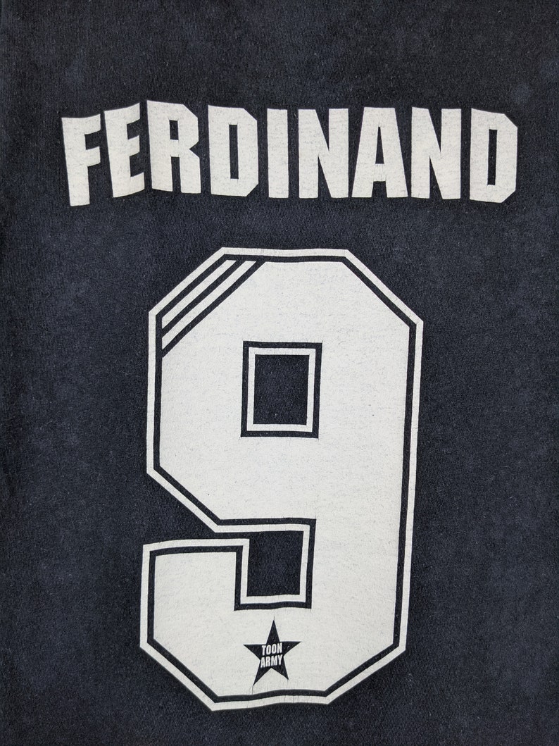 May include: Black t-shirt with white lettering that reads "FERDINAND" and the number "9" in a white block font. A small white star with the text "TOON ARMY" is below the number.