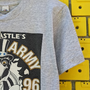 Puede incluir: Camiseta gris con un gr&aacute;fico en blanco y negro de un &aacute;guila de dibujos animados que lleva un sombrero con estrellas y rayas. El gr&aacute;fico tambi&eacute;n incluye el texto "Castle's Army 96".