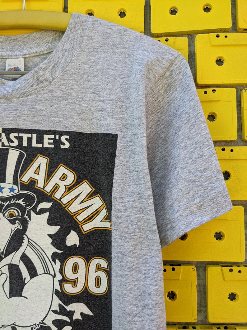Puede incluir: Camiseta gris con un gr&aacute;fico en blanco y negro de un &aacute;guila de dibujos animados que lleva un sombrero con estrellas y rayas. El gr&aacute;fico tambi&eacute;n incluye el texto "Castle's Army 96".
