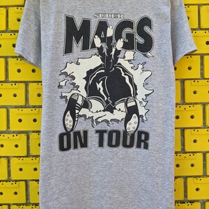 Puede incluir: Camiseta gris con un gr&aacute;fico en blanco y negro de una persona cayendo por un agujero con el texto "SUPER MAGS ON TOUR".
