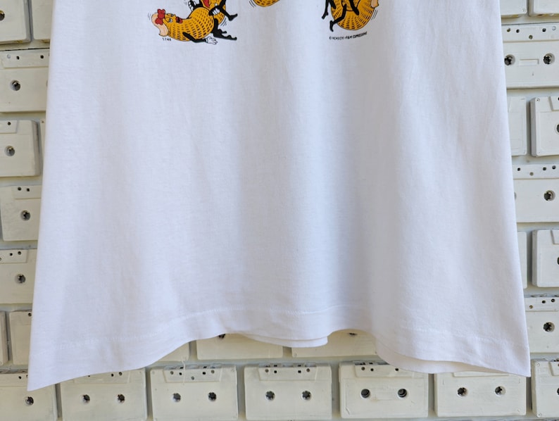 May include: White t-shirt with a cartoon illustration of a group of cats wearing hats and holding a sign that says "It's a cat's life!"