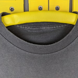 May include: A black t-shirt with a crew neck, hanging on a yellow hanger. The hanger is positioned against a background of yellow cassette tapes.