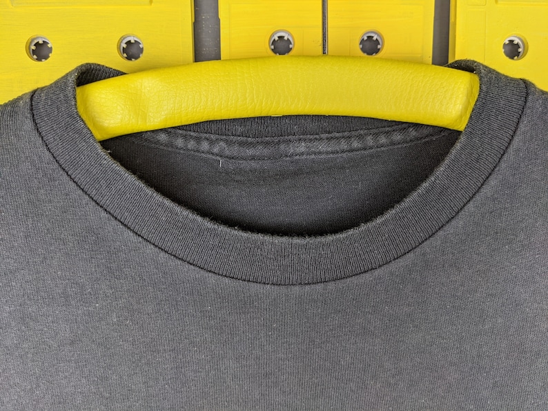 May include: A black t-shirt with a crew neck, hanging on a yellow hanger. The hanger is positioned against a background of yellow cassette tapes.