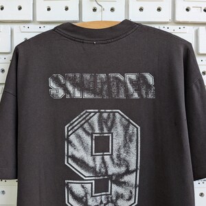 May include: Dark gray t-shirt with the word "SHREDER" printed across the shoulders in a distressed, white font. Below the text is a large number "9" in a tie-dye pattern. The shirt is hanging on a wooden hanger.