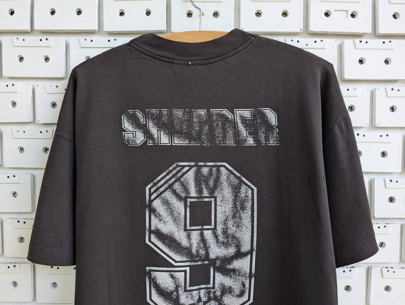 May include: Dark gray t-shirt with the word "SHREDER" printed across the shoulders in a distressed, white font. Below the text is a large number "9" in a tie-dye pattern. The shirt is hanging on a wooden hanger.