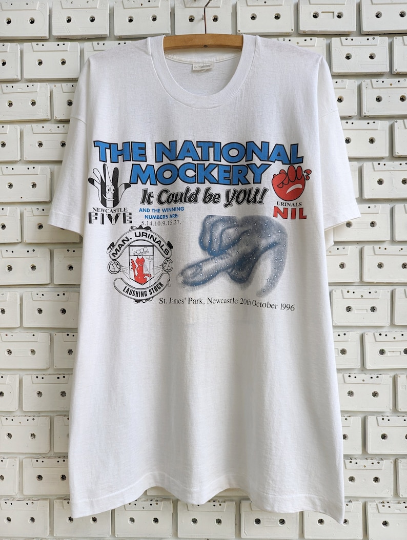 Pu&ograve; includere: Maglietta bianca con una stampa grafica blu e rossa di un calciatore. La grafica include il testo "The National Mockery It Could Be You!" e "Newcastle Five" e "Urinal Nil".