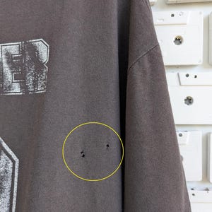 May include: A dark gray t-shirt with a distressed white graphic. The shirt has a crew neck and short sleeves. The fabric shows signs of wear, including small holes. The background features white cassette tapes.