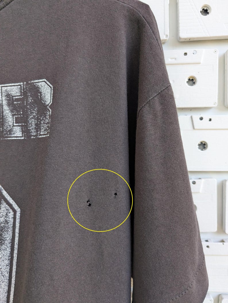May include: A dark gray t-shirt with a distressed white graphic. The shirt has a crew neck and short sleeves. The fabric shows signs of wear, including small holes. The background features white cassette tapes.