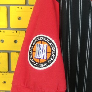 May include: Red and black striped basketball jersey with a red sleeve featuring an embroidered NBA logo patch with the text "National Basketball Association Est. 1946"