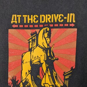 May include: Black t-shirt featuring a yellow and red graphic of a horse pulling a chariot. The text "AT THE DRIVE-IN" is above the graphic and "&iexcl;ATENCI&Oacute;N!" is below the graphic.
