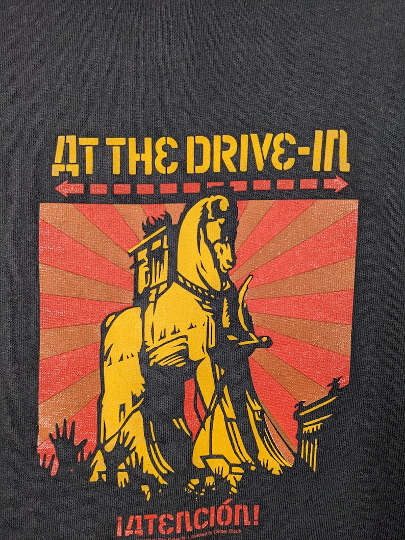 May include: Black t-shirt featuring a yellow and red graphic of a horse pulling a chariot. The text "AT THE DRIVE-IN" is above the graphic and "&iexcl;ATENCI&Oacute;N!" is below the graphic.