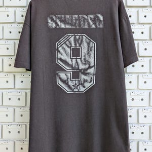 May include: Dark gray t-shirt with the word "SNIDER" printed across the upper back in a distressed, textured font. Below, a large number "9" is displayed in a similar style. The shirt is hanging against a white brick wall.