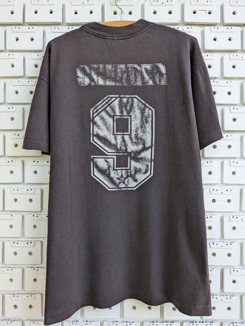 May include: Dark gray t-shirt with the word "SNIDER" printed across the upper back in a distressed, textured font. Below, a large number "9" is displayed in a similar style. The shirt is hanging against a white brick wall.