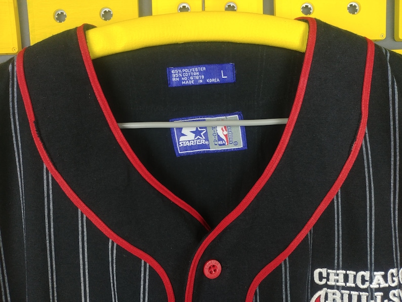 May include: Black and white striped baseball jersey with red trim. The jersey has a red button and a Starter logo with the NBA logo. The text "Chicago Bulls" is embroidered on the front of the jersey.
