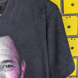 May include: Black t-shirt with a faded image of a man's face on the front. The image is in shades of gray and pink.