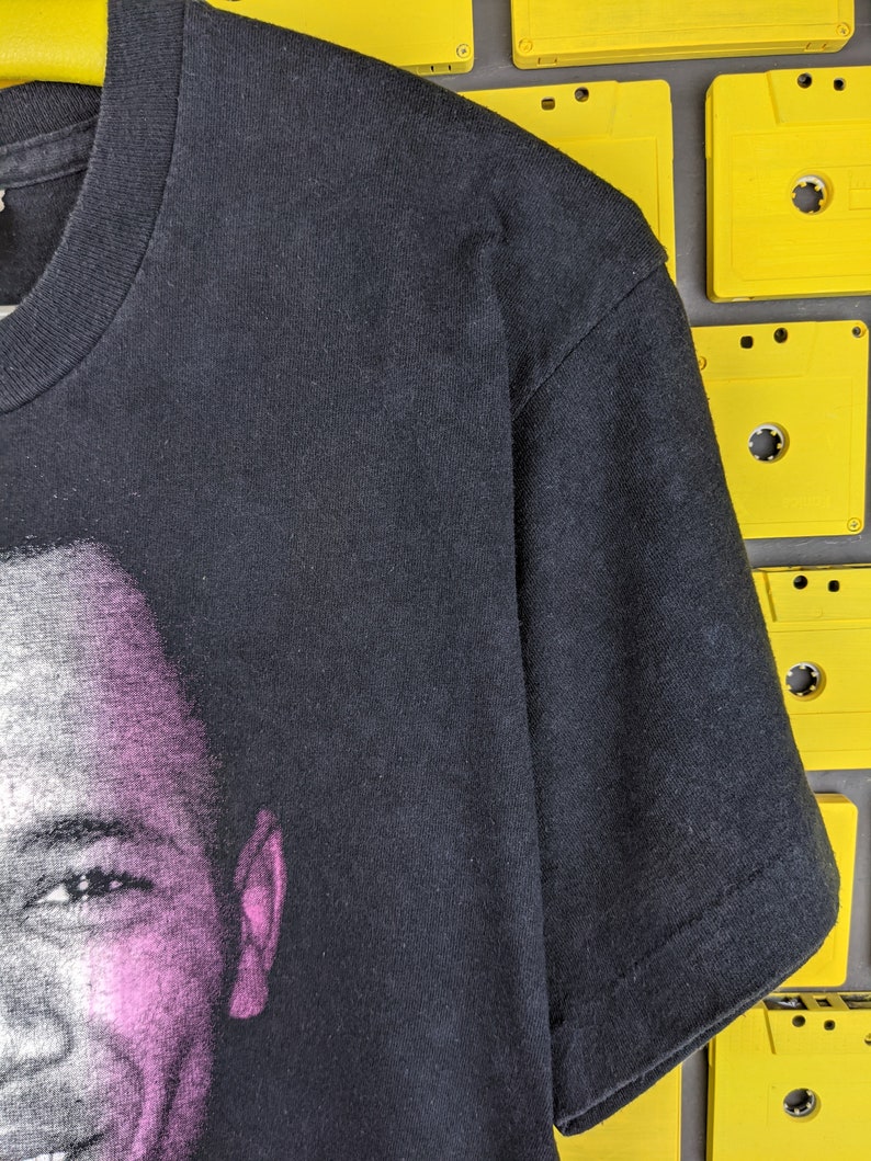 May include: Black t-shirt with a faded image of a man's face on the front. The image is in shades of gray and pink.
