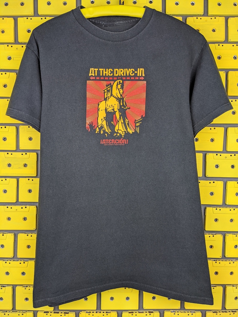 May include: Black t-shirt featuring a graphic of a horse and the text "At The Drive-In" and "&iexcl;ATENCI&Oacute;N!" in red and yellow.