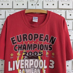 May include: Red t-shirt with the text "EUROPEAN CHAMPIONS 2005 LIVERPOOL 3". The shirt has a crew neck and short sleeves. The graphic includes stars and the years 1977 and 1984.