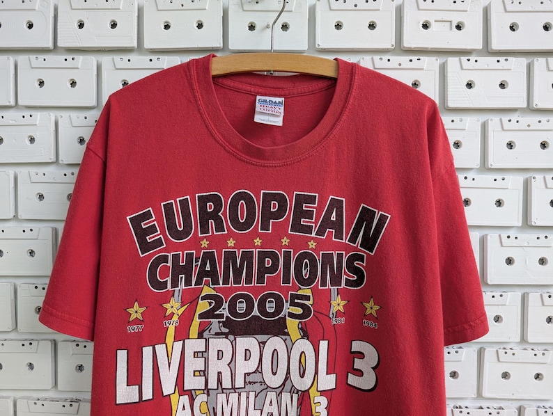 May include: Red t-shirt with the text "EUROPEAN CHAMPIONS 2005 LIVERPOOL 3". The shirt has a crew neck and short sleeves. The graphic includes stars and the years 1977 and 1984.