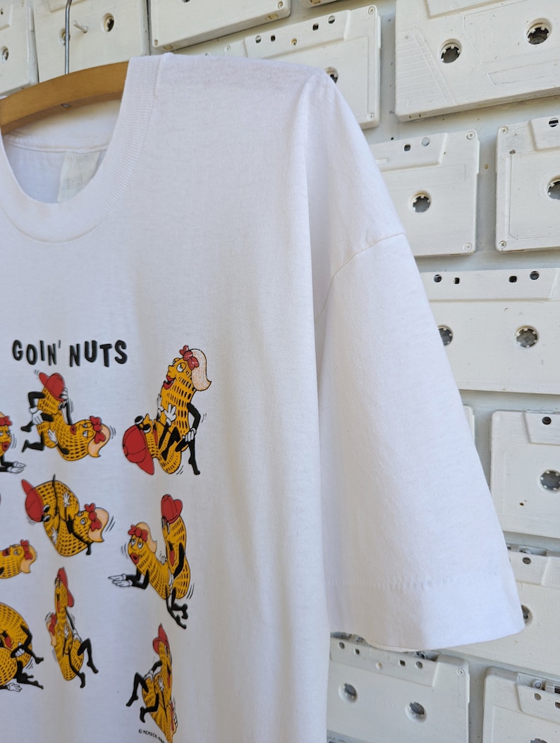 May include: White t-shirt with a graphic of cartoon peanuts in various poses. The text "goin' nuts" is printed at the top of the shirt.